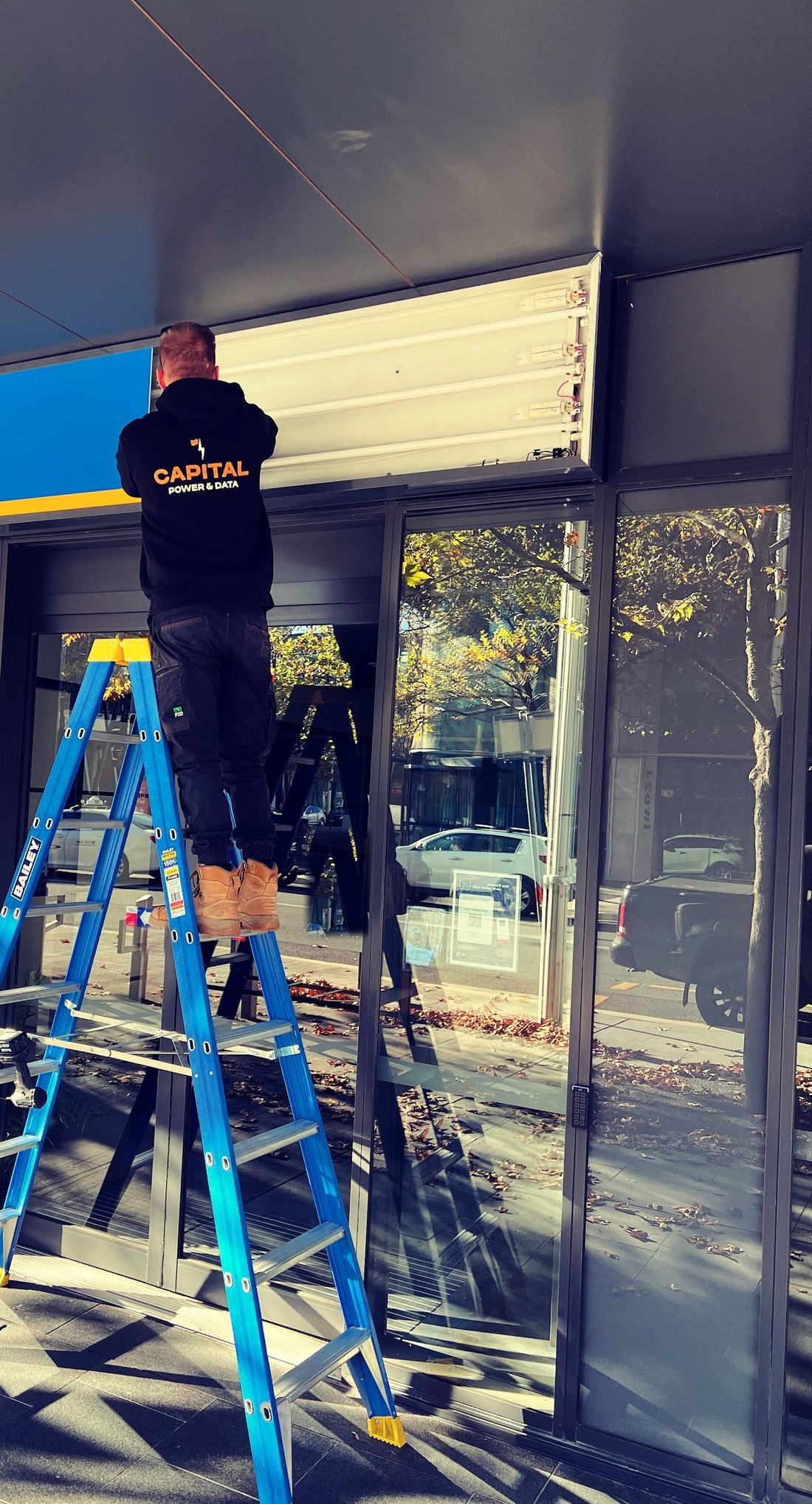 A Capital Power & Data electrician in the branded hoodie up a ladder, working on the lighting above a shopfront